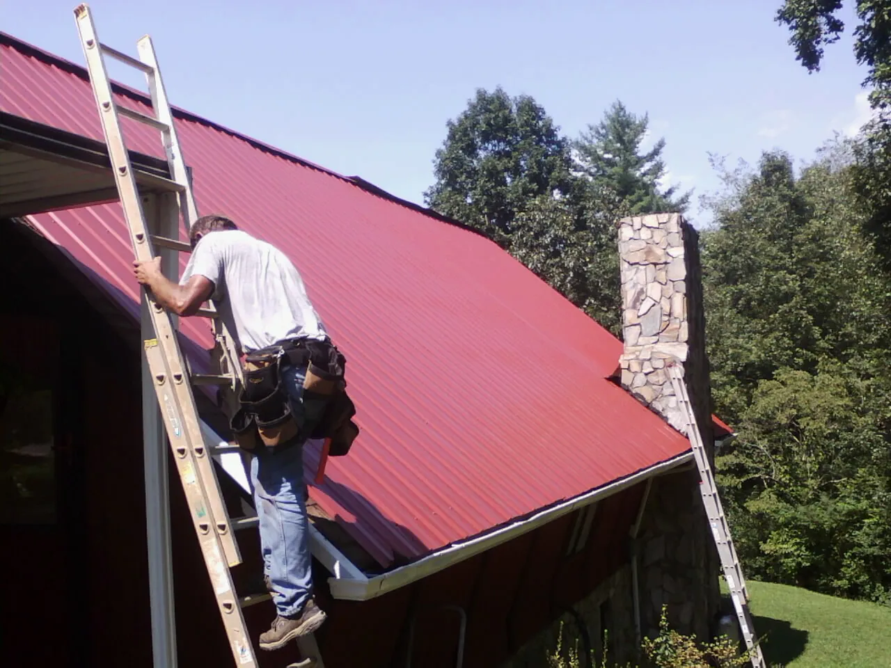 Roofer on a ladder beside a red metal roof and stone chimney — Soffit & Fascia Repair by Roofing Brothers in Odell
